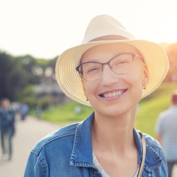Happy young caucasian bald woman in hat and casual clothes enjoying life after surviving breast cancer. Portrait of beautiful hairless girl smiling during walk at city park after curing disease.